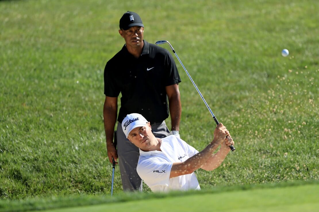 Justin Thomas schlägt einen Ball, Tiger Woods schaut im Hintergrund zu.