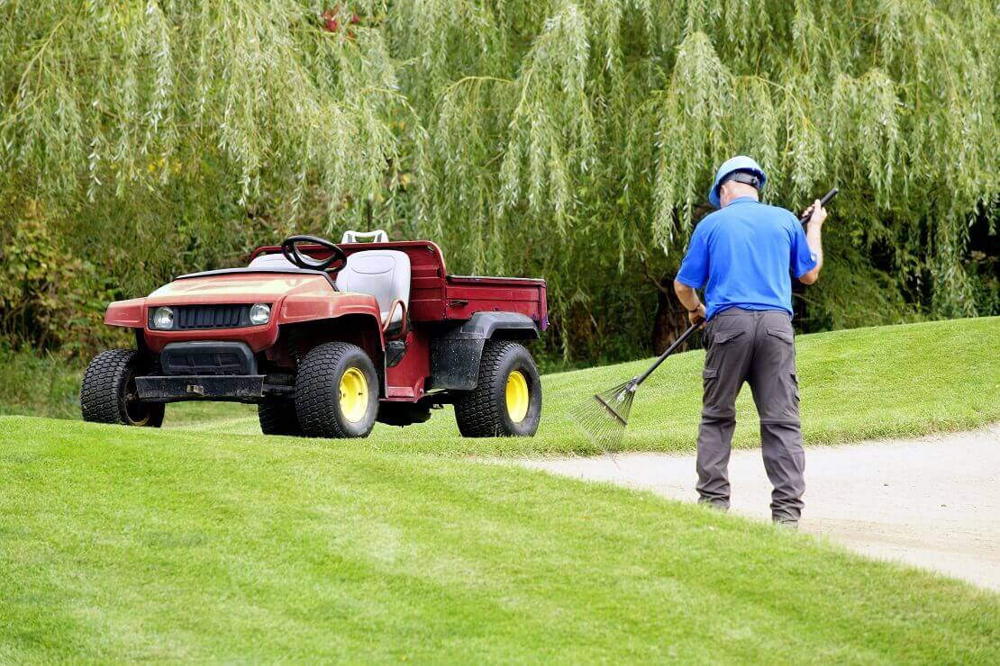 Ein Greenkeeper hakt den Sand eines Bunkers und vor ihm steht sein Golfcart.