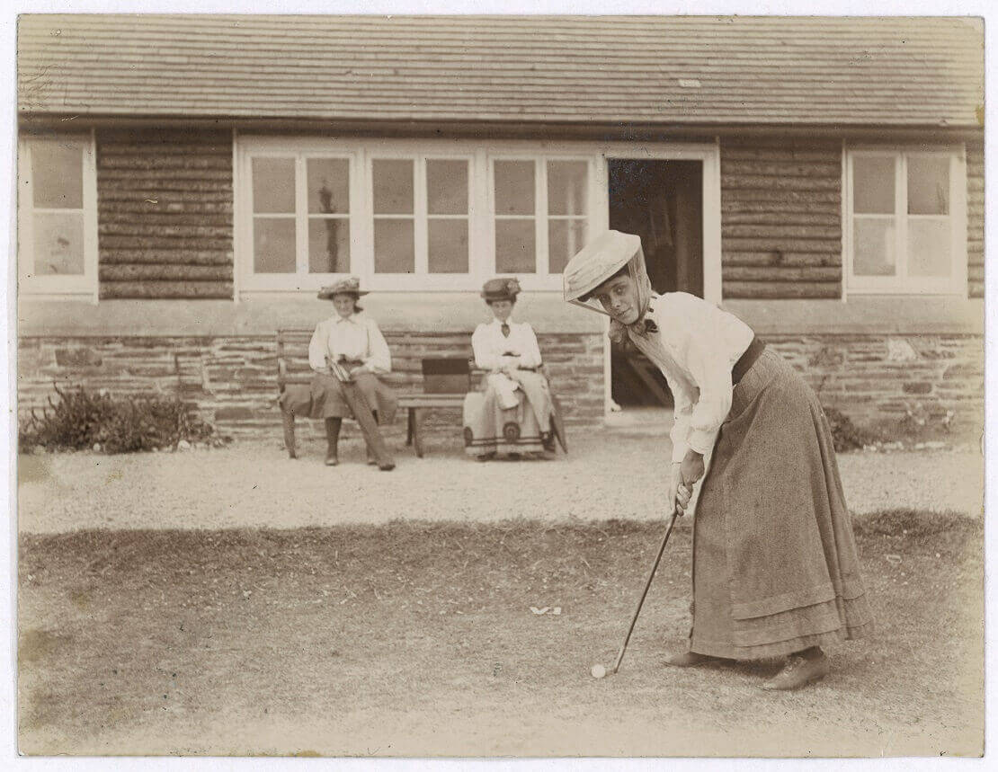 Altes Bild von einer Frau, die am Abschlag steht und zwei weitere Frauen sitzen im Hintergrund auf einer Bank und schauen zu.
