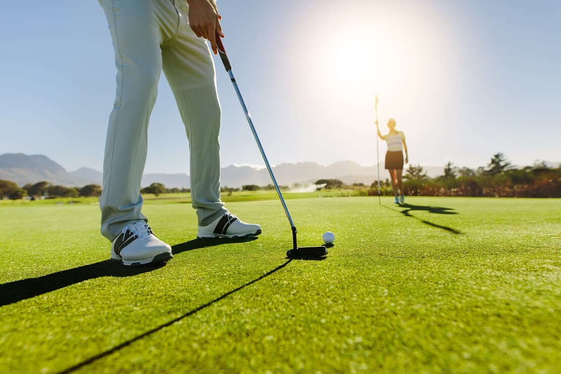 Ein Golfer schlägt im Vordergrund einen Golfball ans Loch an der Fahne steht eine Golferin und wartet