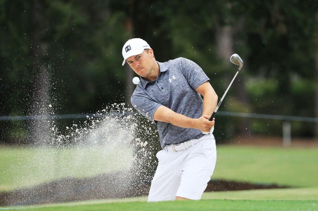 Jordan Spieth schlägt den Ball aus einem Sandbunker und Sand fliegt mit dem Ball.