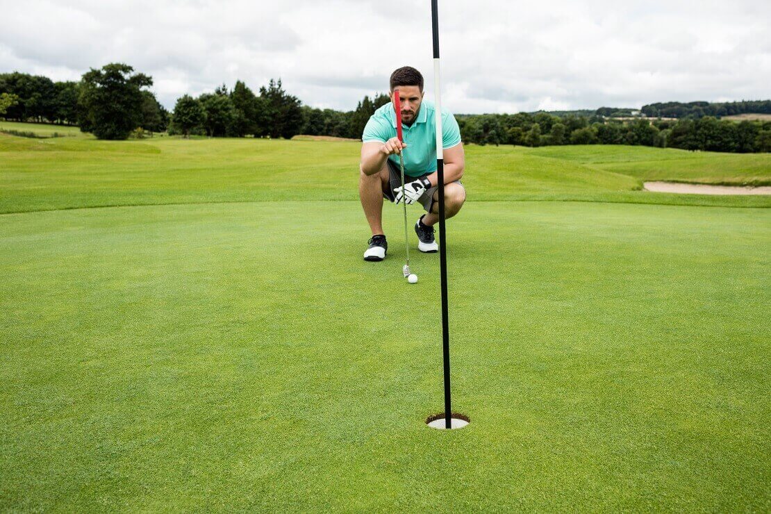 Im Vordergrund das Loch mit Fahne und im Hintergrund ein Golfer, der in der Hocke mit Ball und Schläger das Grün liest.