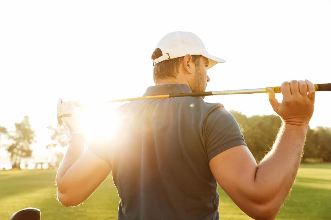 Golfer steht mit dem Rücken zum Betrachter und hält seinen Schläger hinter seinen Rücken während die Sonne von vorne scheint.