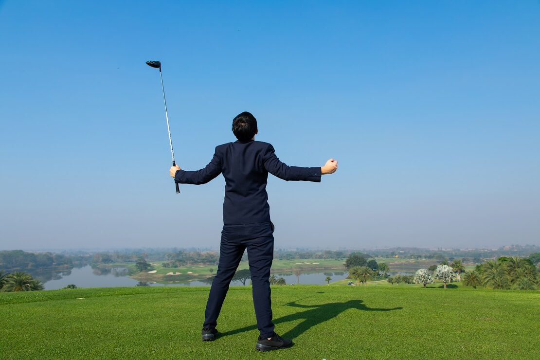 Geschäftsmann hält den Schläger in der Hand und macht eine freiheitsausdrückende Pose auf dem Golfplatz