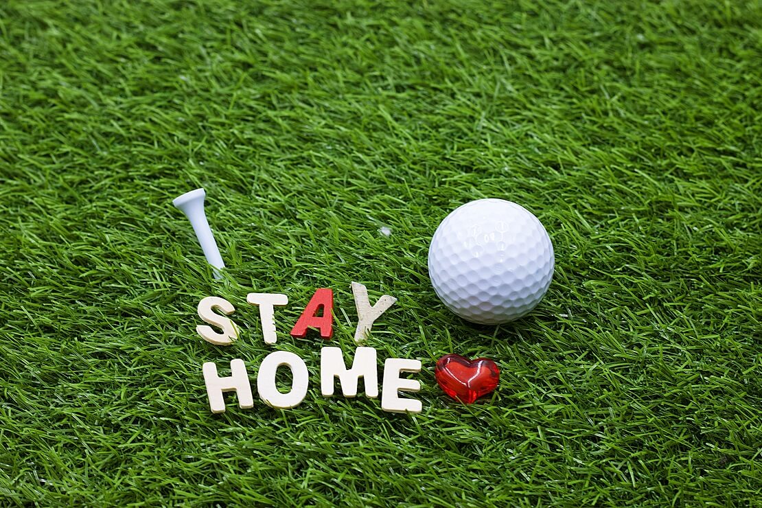 "Stay Home" aus einzelnen Buchstaben, Tee und Golfball liegen auf grünem Rasen