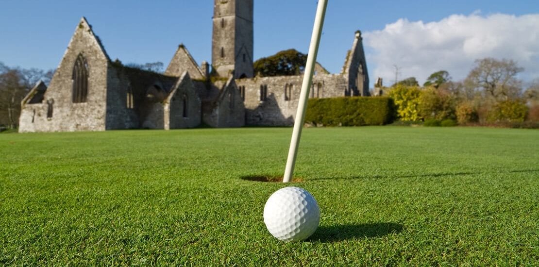 Golfball am Loch eines irischen Golfclubs mit einer Abtei im Hinzergrund
