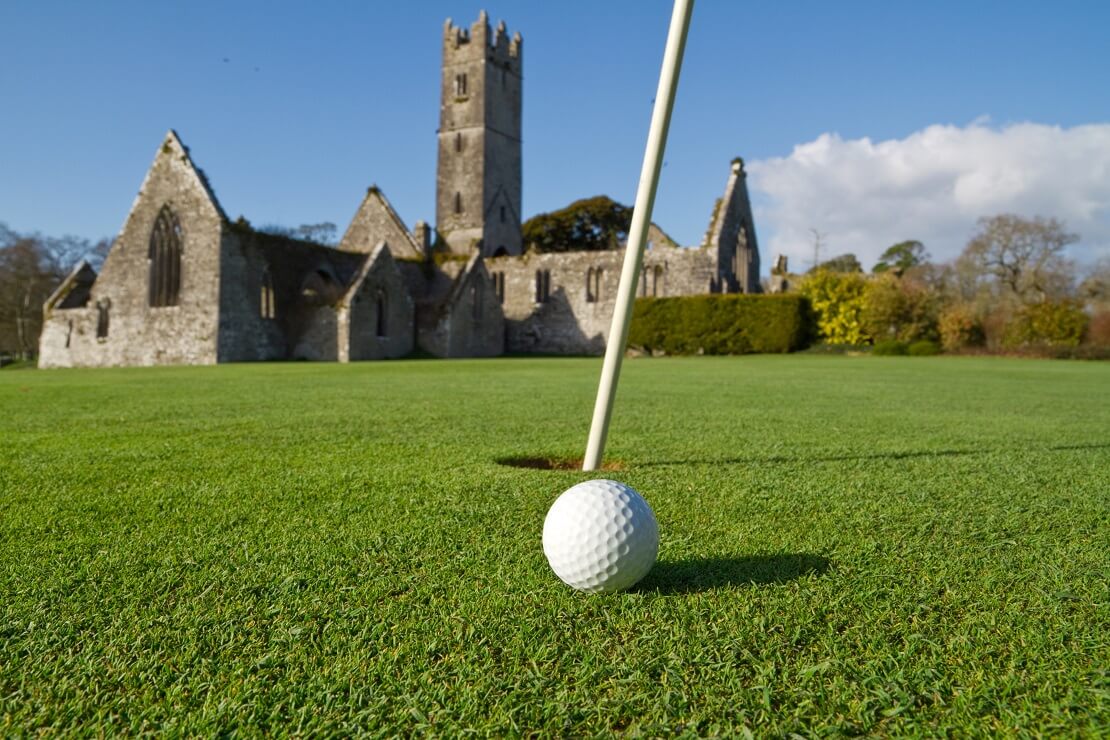 Golfball am Loch eines irischen Golfclubs mit einer Abtei im Hinzergrund