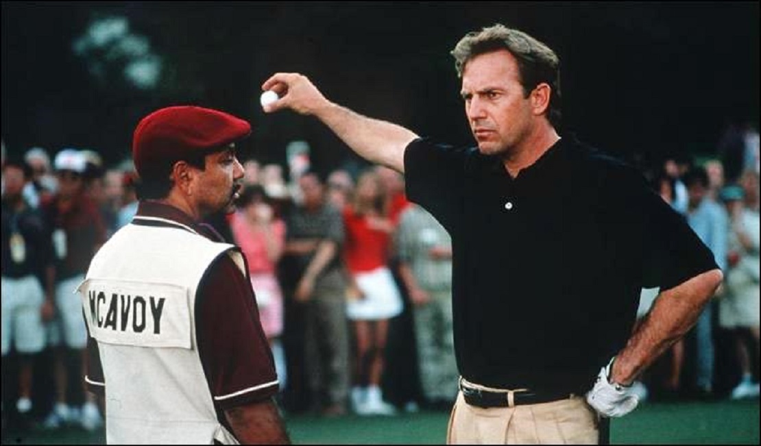 Kevin Costner steht vor seinem Caddy, streckt den Arm aus und lässt den Golfball fallen
