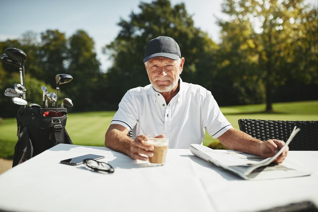 Senior sitzt am Tisch einer Golfanlage und trinkt Kaffee