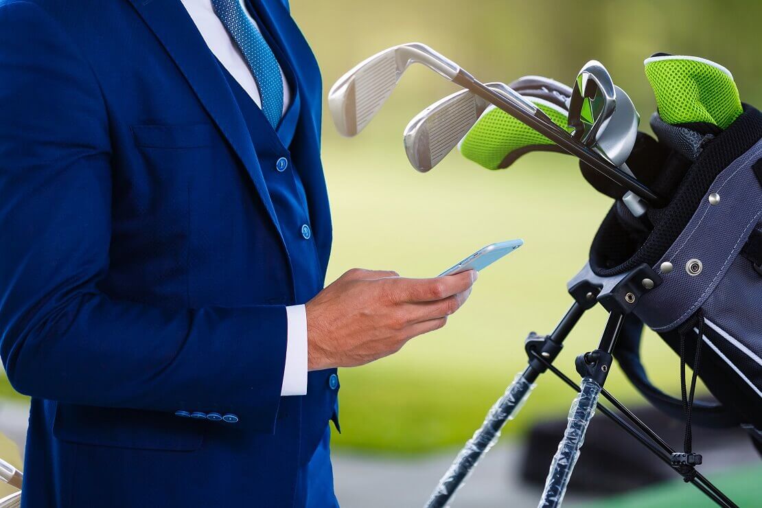Geschäftsmann im Anzug mit Handy in der Hand und Golf-Equipment