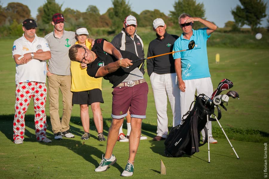 Sieben Golfer stehen in einem Halbkreis, einer Schlägt den Ball.