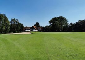 Ein leicht unebener Golfpark mit einem Landhaus und Bäumen außen rum.