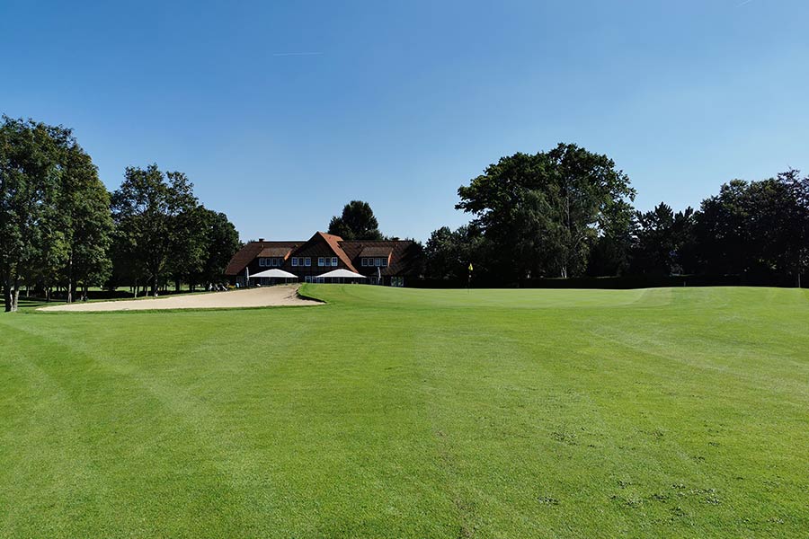 Ein leicht unebener Golfpark mit einem Landhaus und Bäumen außen rum.