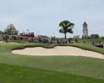 Golfer auf einem hügeligen Golfplatz mit Sandbunkern, im Hintergrund Fans und ein Kirchturm