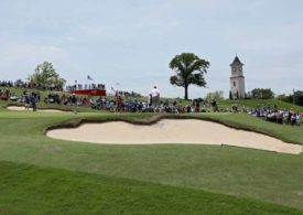 Golfer auf einem hügeligen Golfplatz mit Sandbunkern, im Hintergrund Fans und ein Kirchturm