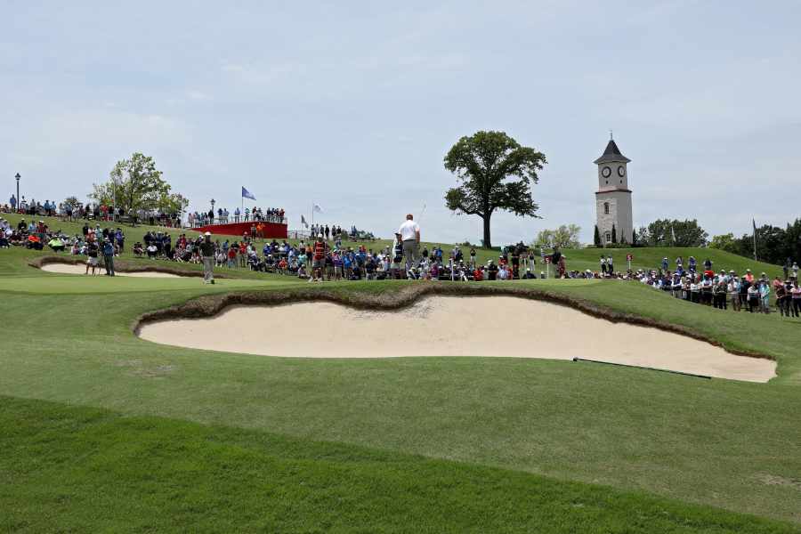 Golfer auf einem hügeligen Golfplatz mit Sandbunkern, im Hintergrund Fans und ein Kirchturm