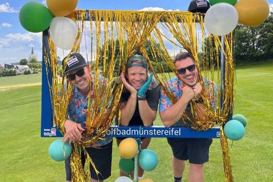 Drei Leute stehen in einem geschmücktem Fotorahmen auf dem Golfplatz