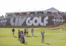 Ein Golfer mit Caddie und Kamerateam vor einer Zuschauer-Tribüne