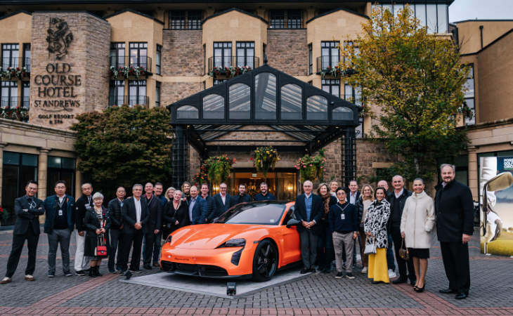 Eine Gruppe Menschen vor einem schönen Hotelgebäude, in der Mitte ein oranger Porsche
