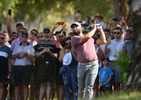 Jon Rahm steht im Vordergrund und schwingt sein Golfschläger und im Hintergrund stehen viele Zuschauer die ihn filmen.