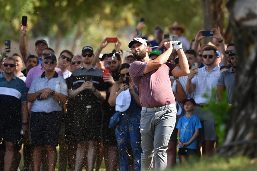 Jon Rahm steht im Vordergrund und schwingt sein Golfschläger und im Hintergrund stehen viele Zuschauer die ihn filmen.