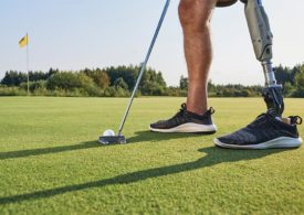 Ein Bild wo man ein Golfschläger, Golfball und Beine von einem Mann sieht und sein linkes Bein ist eine Prothese
