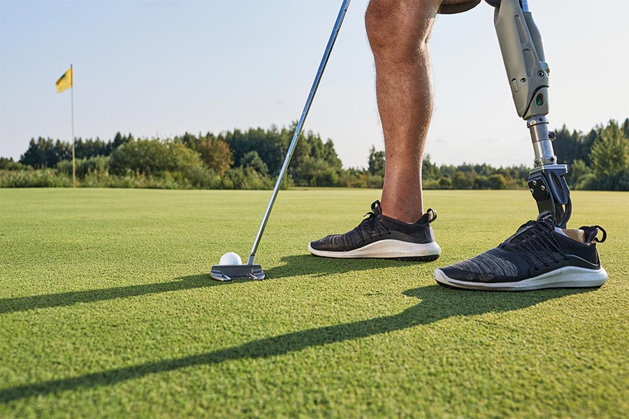 Ein Bild wo man ein Golfschläger, Golfball und Beine von einem Mann sieht und sein linkes Bein ist eine Prothese