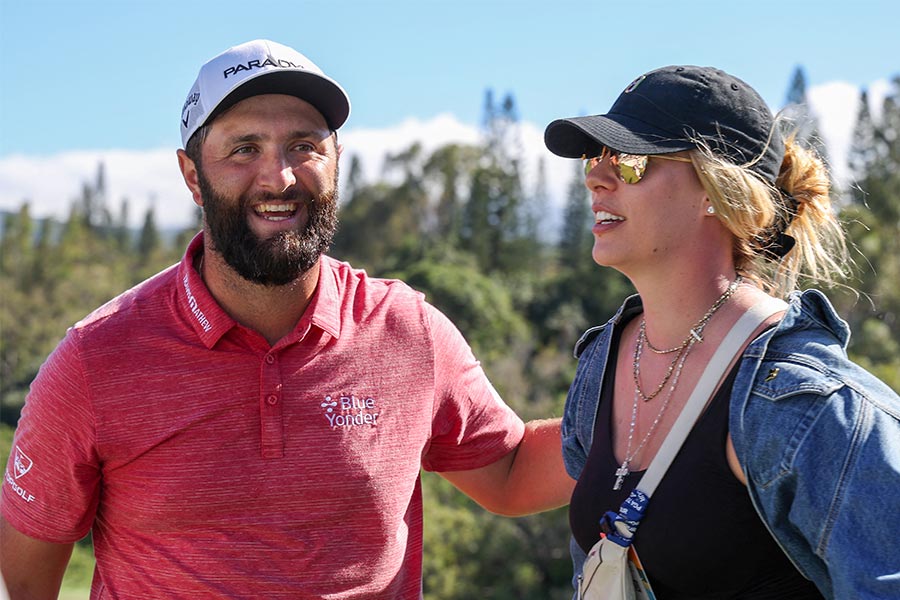 Links steht ein männlich Golfspieler in einem roten T-Shirt und rechts steht eine Frau in einer Jeans Jacke und hat eine Sonnenbrille auf