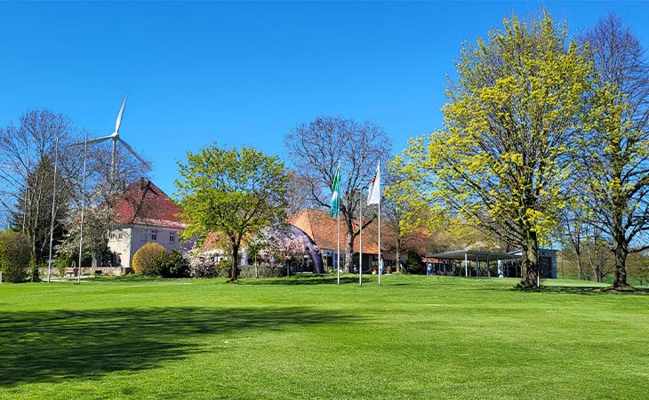 Ein grüner Rasen mit drei Flaggen in der Mitte und ganz hinten sieht man zwei Gebäude