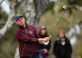 Ein Golfer in einer roten Sportjacke schwingt ein Golfschläger und man sieht zwei Personen im Hintergrund