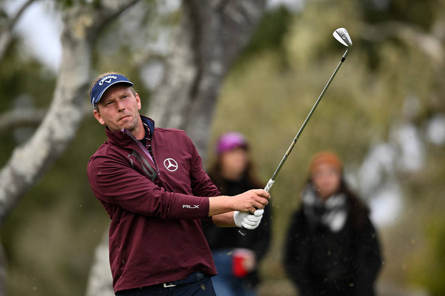 Ein Golfer in einer roten Sportjacke schwingt ein Golfschläger und man sieht zwei Personen im Hintergrund