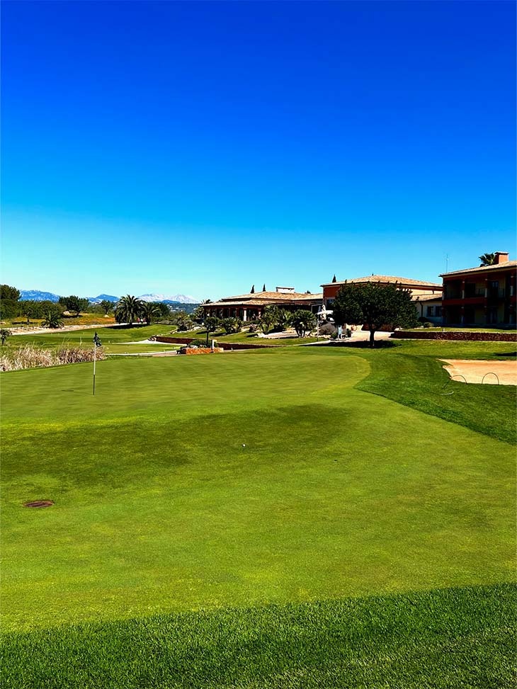 Ein Golfplatz mit einer Flagge in der Mitte und rechts im Hintergrund stehen mehrere Gebäude