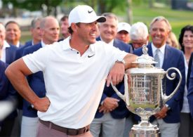 Brooks Koepka stützt sich auf einen silbernen Pokal ab und im Hintergrund stehen mehrere Menschen im blauen Jackett