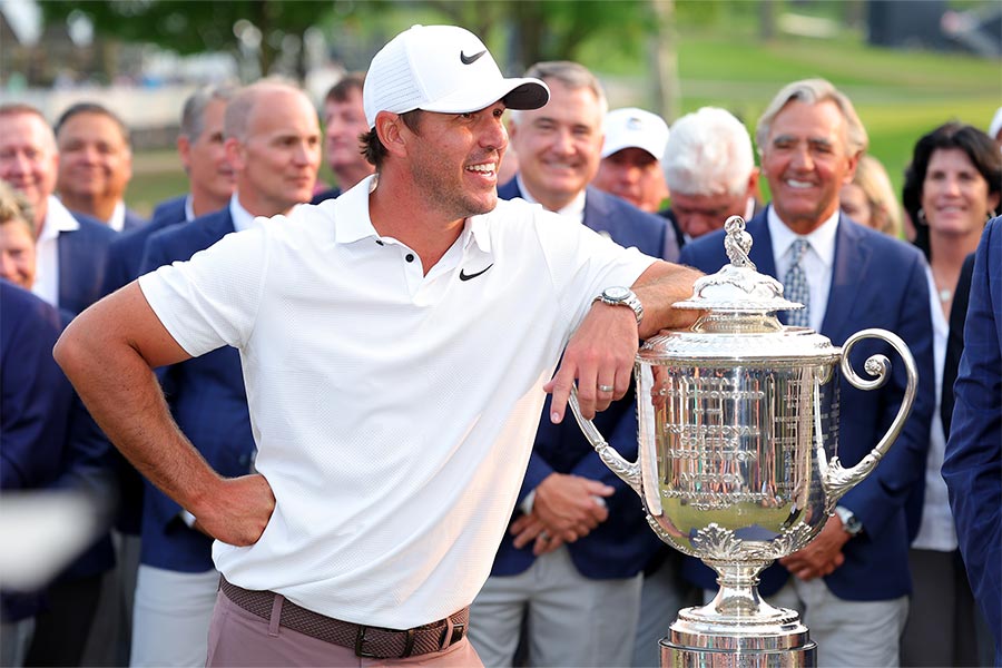 Brooks Koepka stützt sich auf einen silbernen Pokal ab und im Hintergrund stehen mehrere Menschen im blauen Jackett