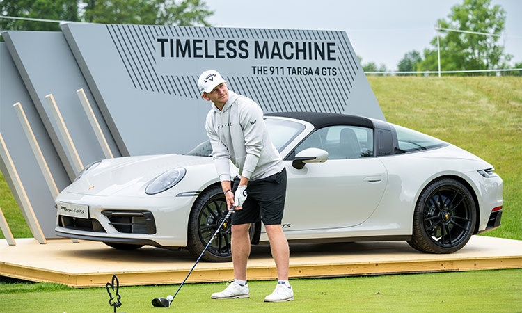 Der Golfspieler Nick Bachem steht mit seinem Golfschläger vor einem grauen Porsche