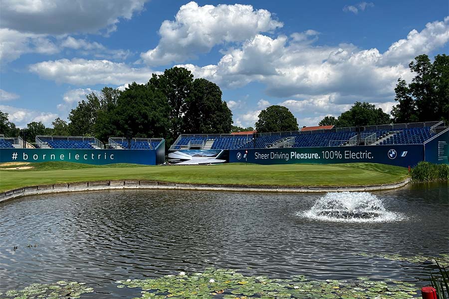 Ein Teich mit einer Wasserfontäne und im Hintergrund ist ein Golfplatz mit einer Tribüne