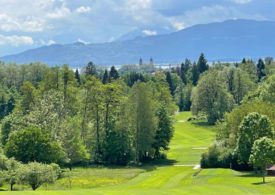 Blick über einen Golfplatz, im Hintergrund ein Kirchturm und Berge