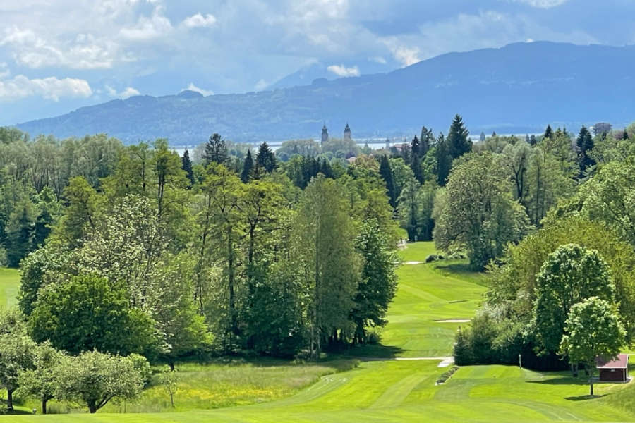 Blick über einen Golfplatz, im Hintergrund ein Kirchturm und Berge