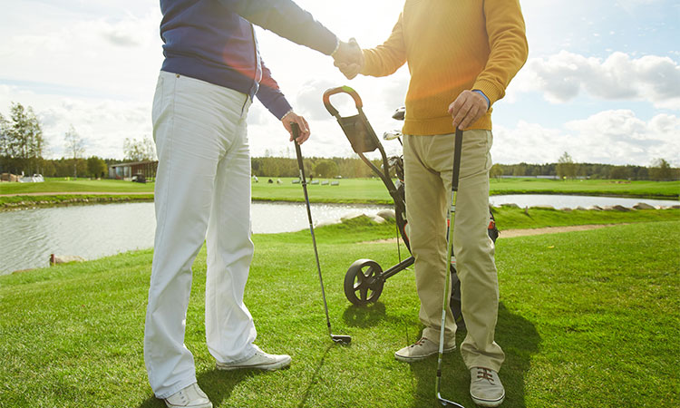 Zwei Golfer geben sich die Hand auf einem Golf Feld und halten dabei beide jeweils ein Golfschläger in der anderen Hand
