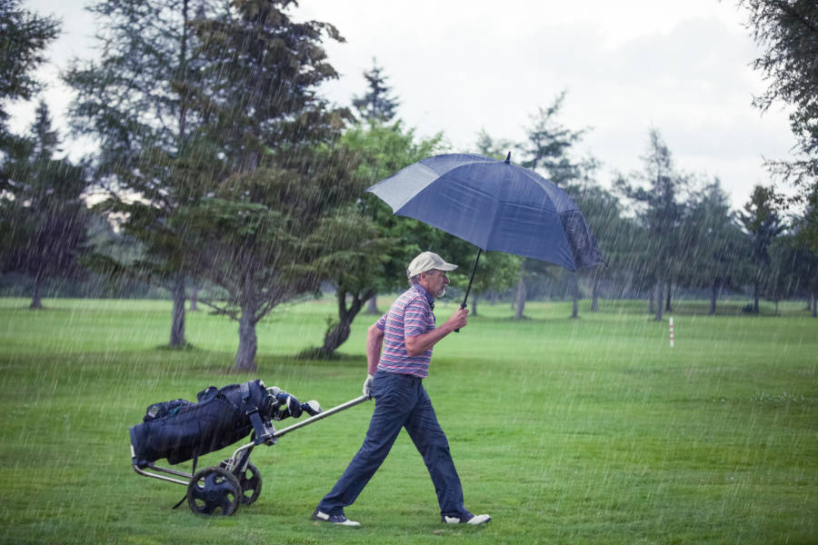 Ein Golfer hat einen Schirm in der Hand und zieht im Regen seinen Trolley hinterher