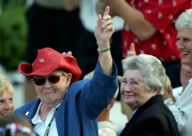 Shirley Spork mit rotem Hut reckt den Zeigefinger in die Luft, daneben steht Louise Suggs