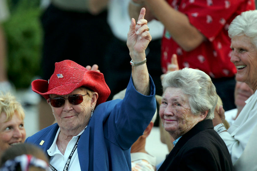 Shirley Spork mit rotem Hut reckt den Zeigefinger in die Luft, daneben steht Louise Suggs