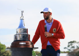 Scottie Scheffler in der roten Jacke des Arnold Palmer Invitational spricht mit einem Mikrofon in der Hand vor der Trophäe